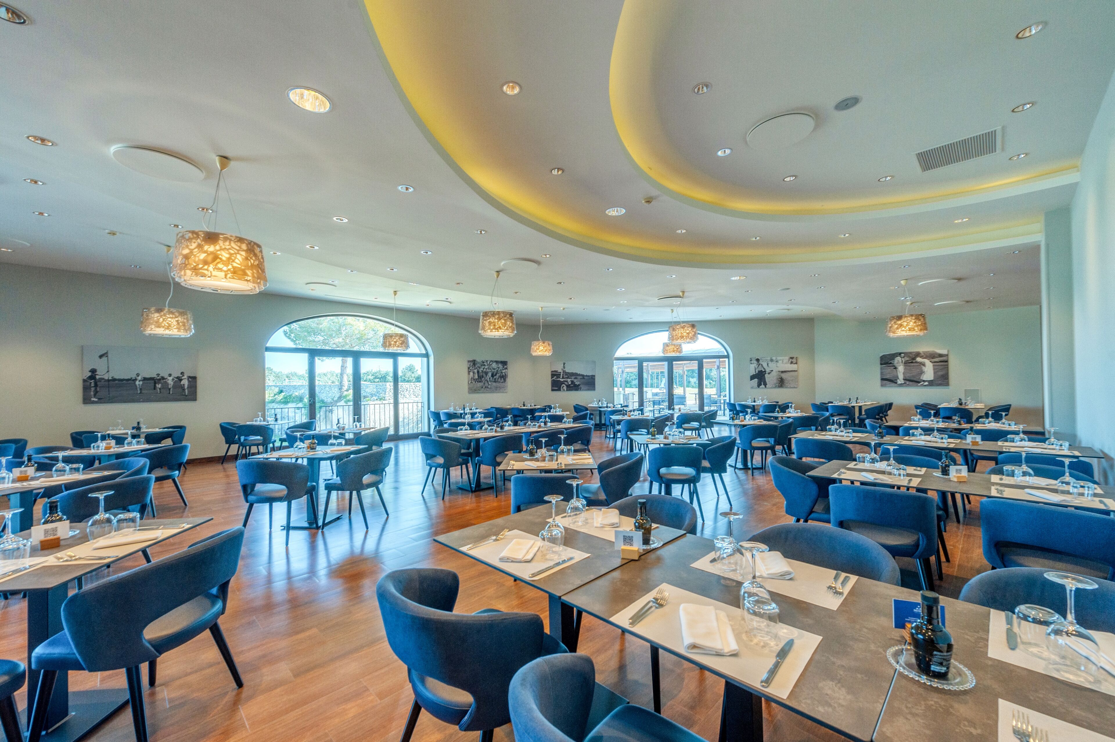 A spacious restaurant with multiple tables and chairs, large windows, and modern lighting fixtures.