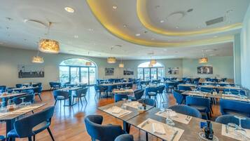 A spacious restaurant with multiple tables and chairs, large windows, and modern lighting fixtures.