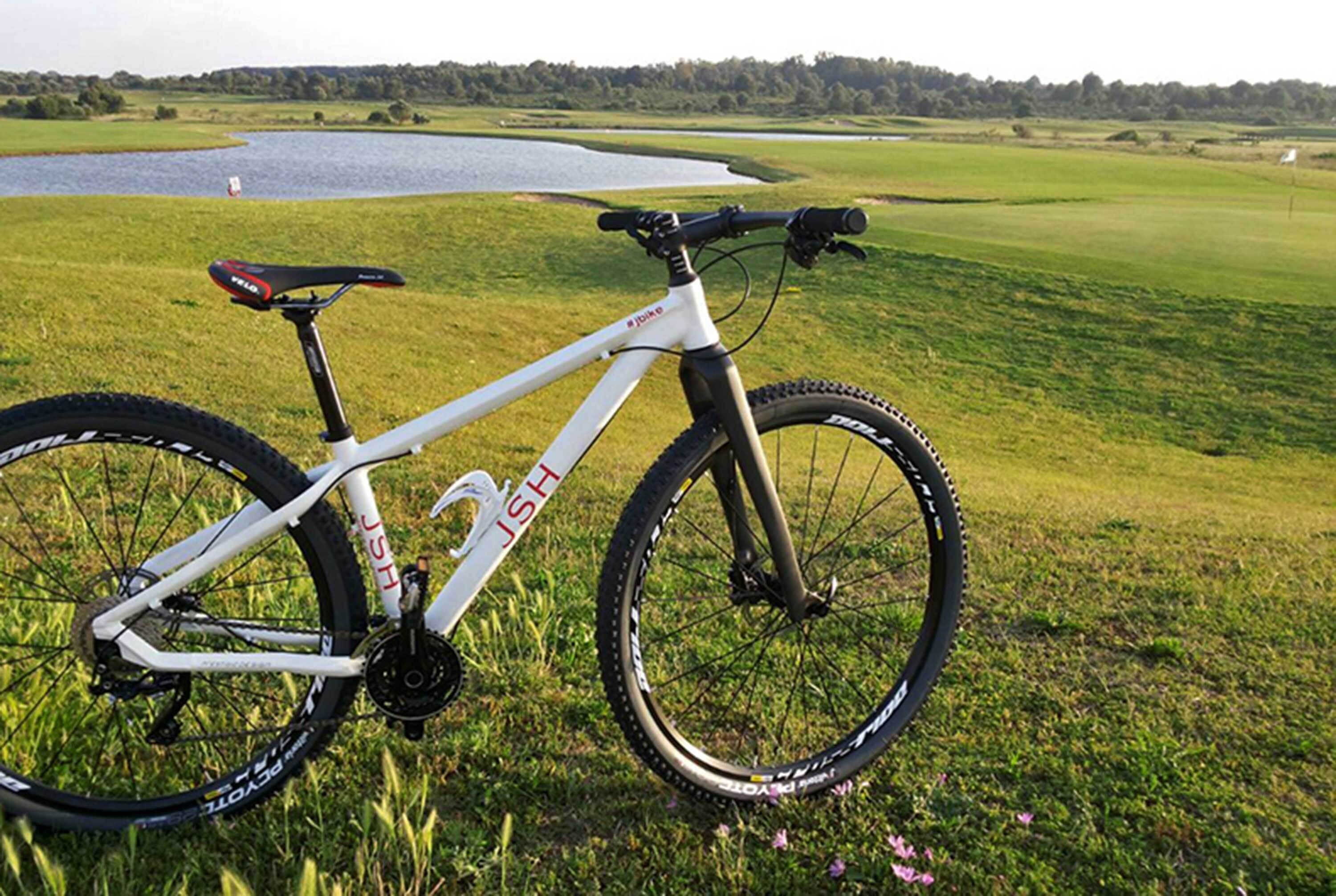 Una bicicleta de montaña blanca con el nombre de marca 'JSH' en el cuadro, estacionada sobre césped con un lago y un campo de golf al fondo.