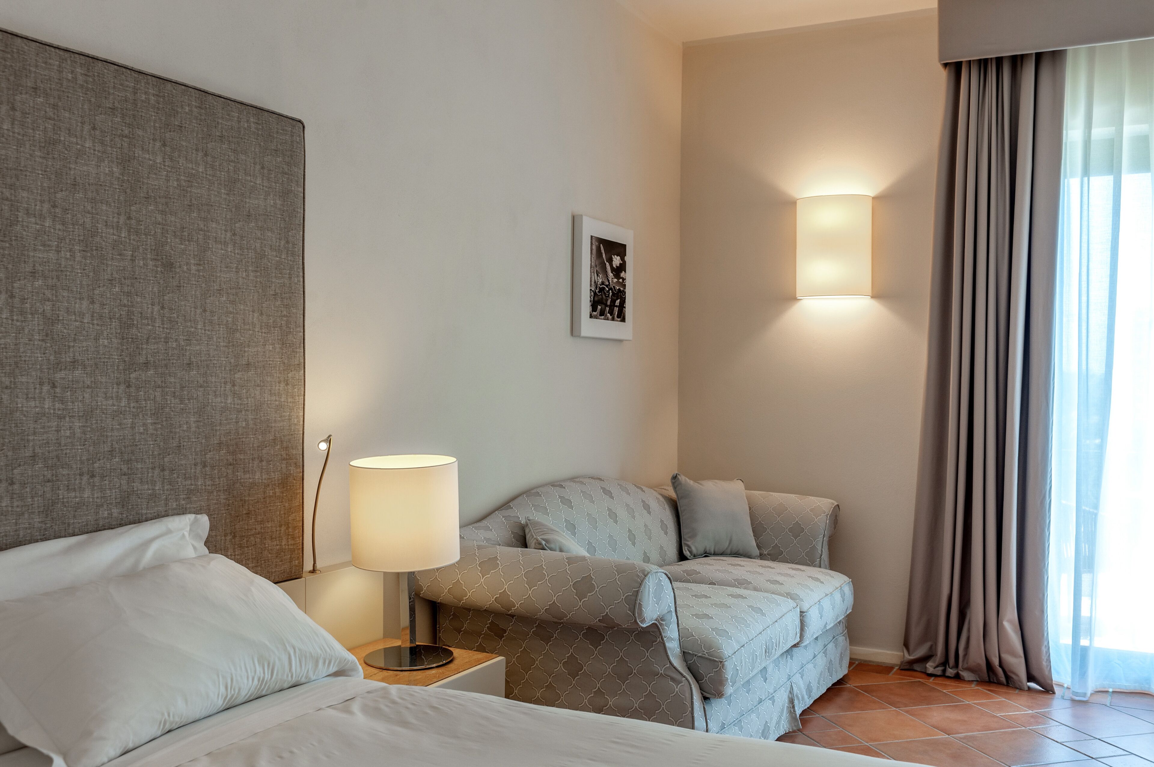 A hotel room with a bed, a nightstand with a lamp, a sofa, and a wall-mounted light fixture.