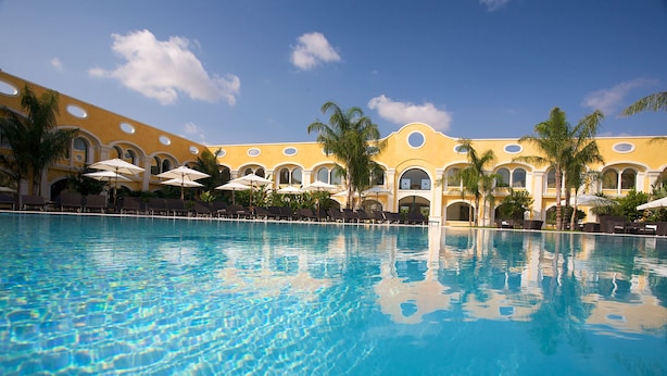 A pool with clear blue water, surrounded by palm trees and a yellow building with arched windows. - Acaya Golf Resort & SPA (Vernole)