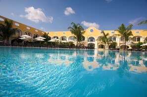 A pool with clear blue water, surrounded by palm trees and a yellow building with arched windows. - Acaya Golf Resort & SPA (Vernole)