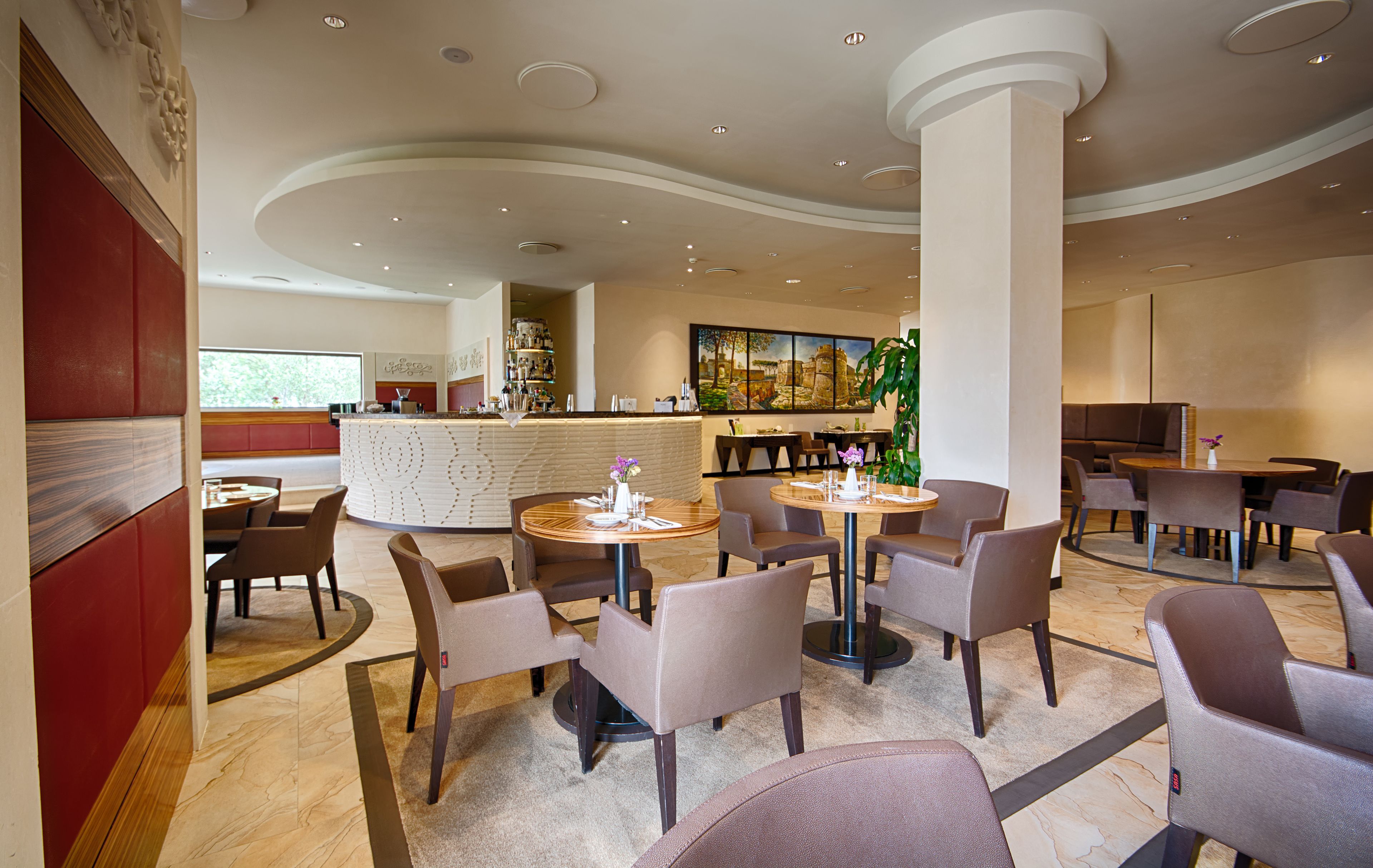 Eine moderne Hotelhalle mit runden Tischen, bequemen Sitzgelegenheiten, einer Bar und einem großen Gemälde an der Wand.