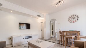 A modern living room with a flat-screen TV, a white coffee table, and a dining area with wooden chairs and a table. - Puerto Banús Luxury Penthouse (Marbella)