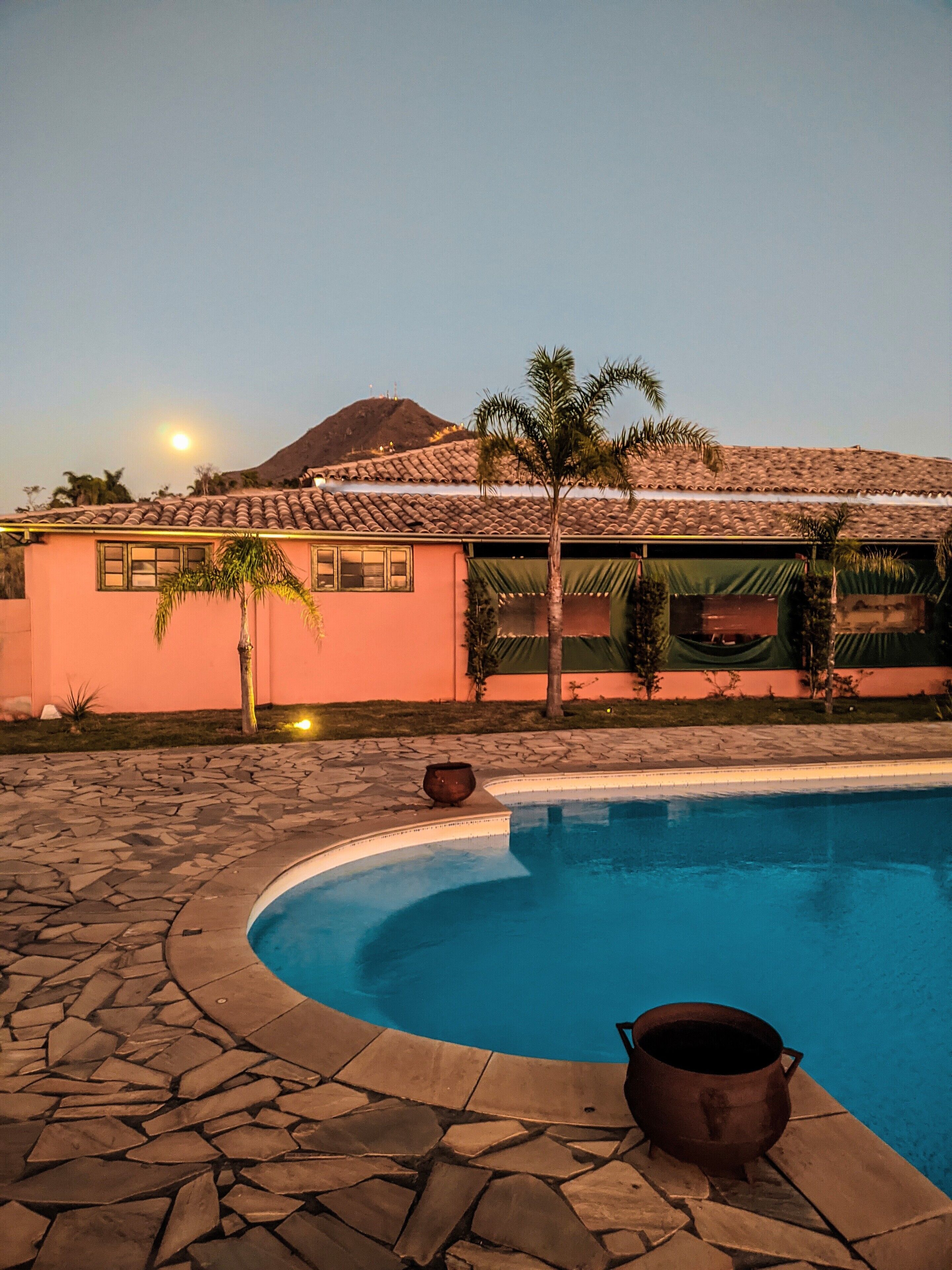 A swimming pool with stone tiles, a building with a tiled roof, and a mountain in the background.
