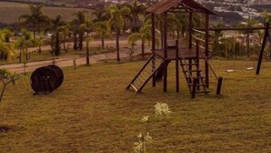 A playground with a swing set and a slide, surrounded by grass and palm trees, with a sunset in the background. - Hotel Varandas da Montanha (Carmo do Rio Claro)