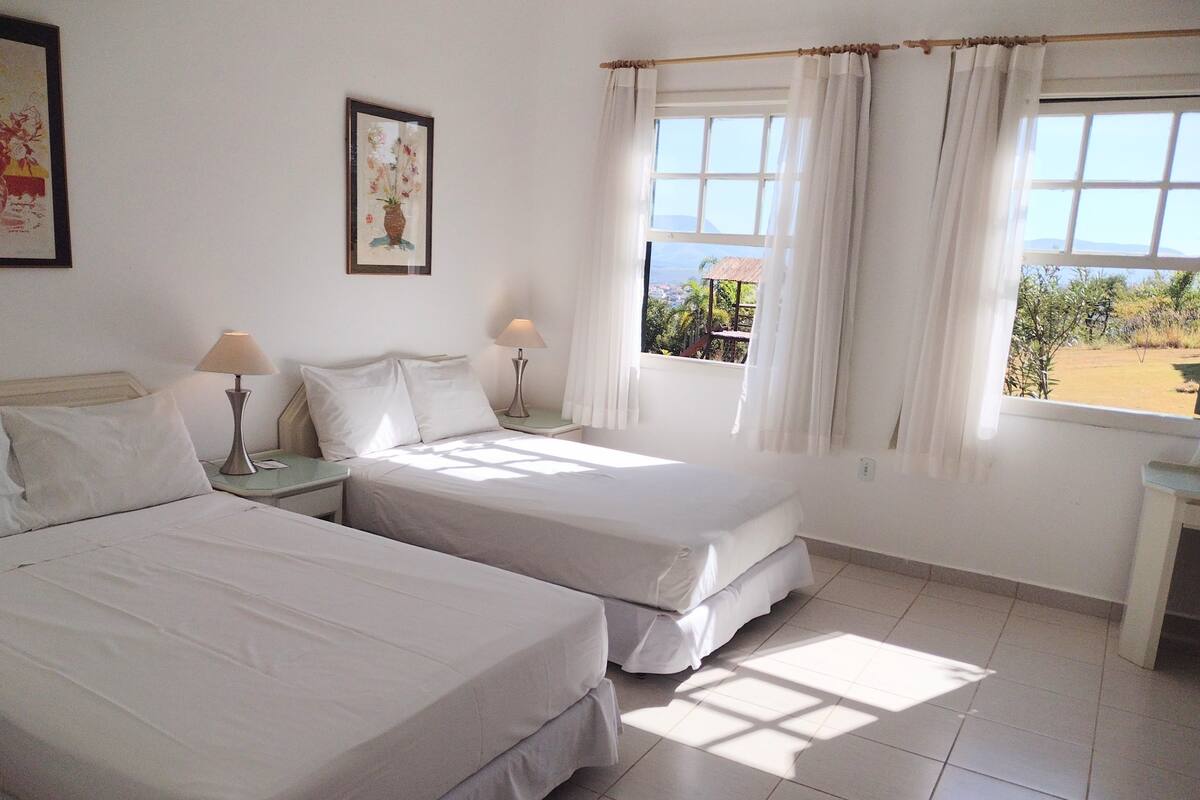 A room with two beds, a window with curtains, a framed picture on the wall, and a small table.