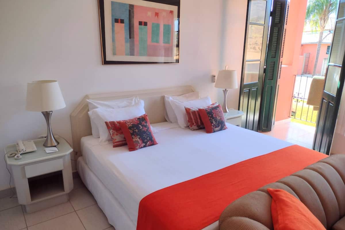 A hotel room with a bed, two bedside tables, a lamp, a painting, and a view of the outside.