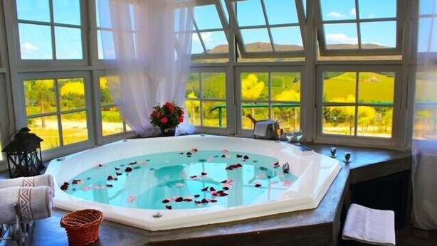 A hot tub with rose petals floating on the water, surrounded by large windows with a view of green fields. - Hotel Varandas da Montanha (Carmo do Rio Claro)