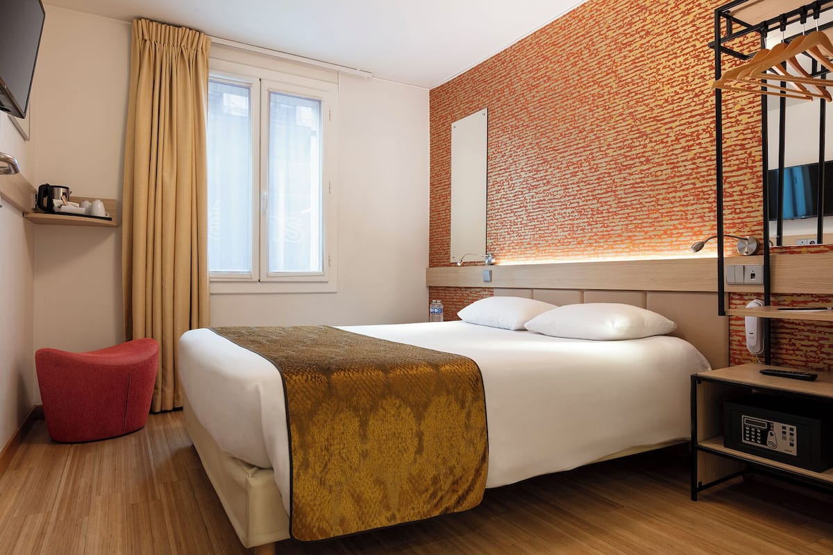 Een hotelkamer met een bed, een rode stoel, een houten vloer, een raam met gordijnen, een muur met een baksteenmotief, een spiegel en een plank met een afstandsbediening.