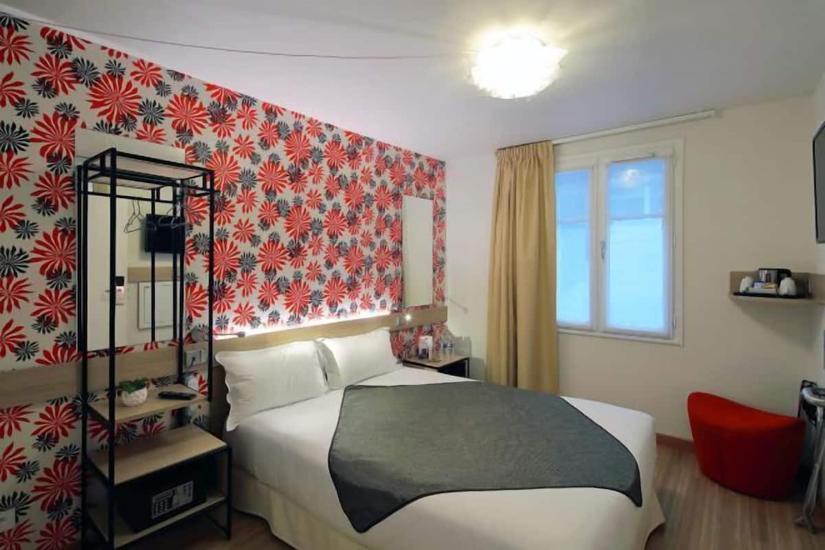 Een hotelkamer met een bed, nachtkastje, spiegel, kledingkast en een raam met gordijnen.