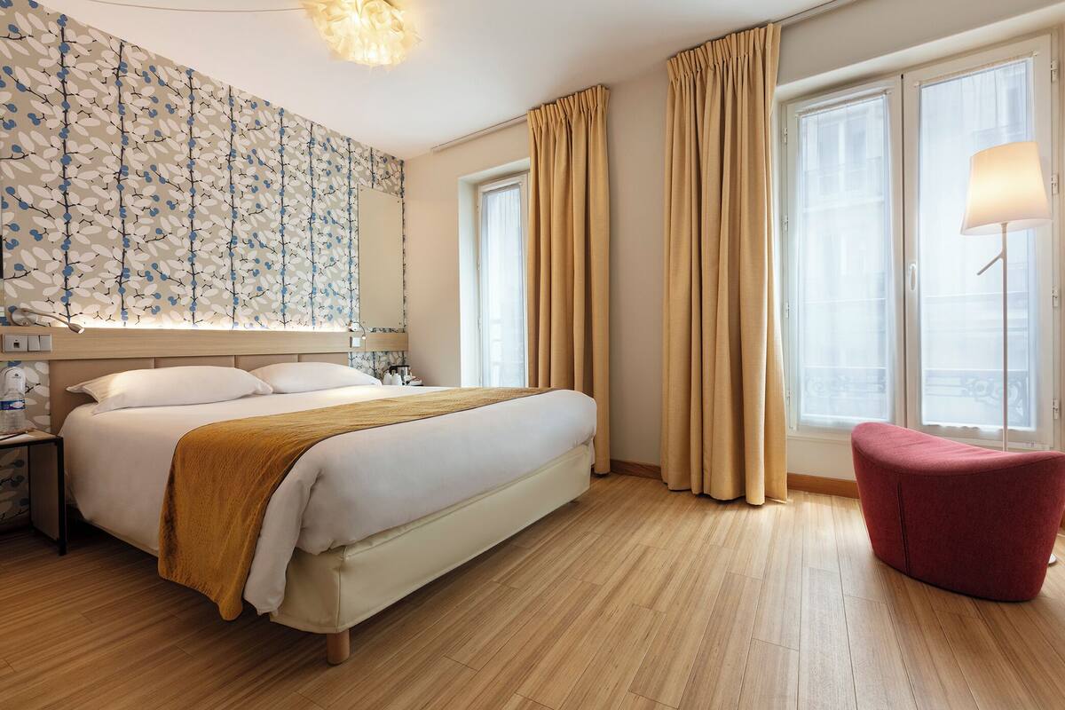 Een hotelkamer met een groot bed, een nachtkastje, een rode stoel en een raam met gordijnen.
