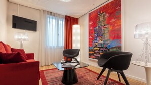 Un salon moderne avec un canapé rouge, des fauteuils noirs, une table basse en verre et une fresque colorée sur le mur.