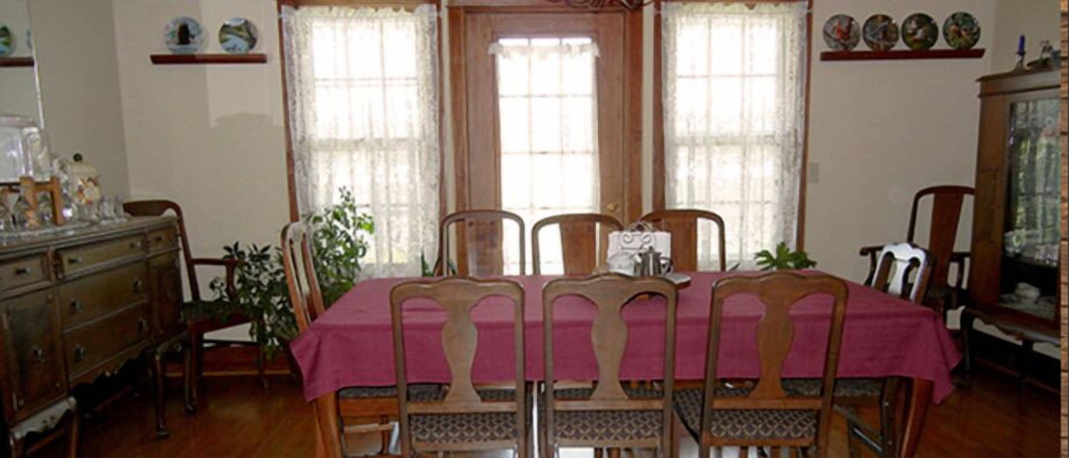Una sala da pranzo con un tavolo in legno apparecchiato per quattro persone, ricoperto da una tovaglia rosa e circondato da sedie in legno con decorazioni elaborate sulla schienale.