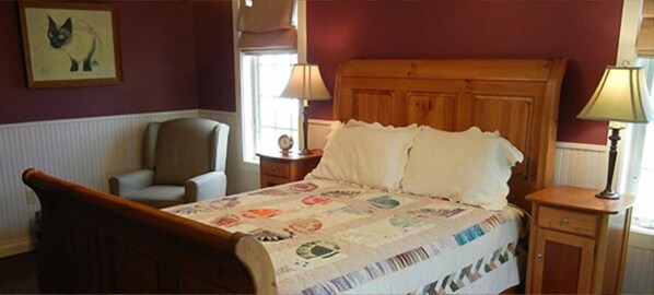 Un dormitorio con una cama de madera, una silla, una mesita de noche con una lámpara y un cuadro enmarcado en la pared.