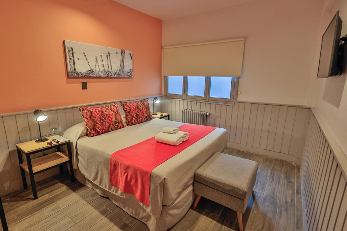 Une chambre à coucher comprenant un lit, des tables de chevet, un banc, un radiateur et une fenêtre avec des stores.
