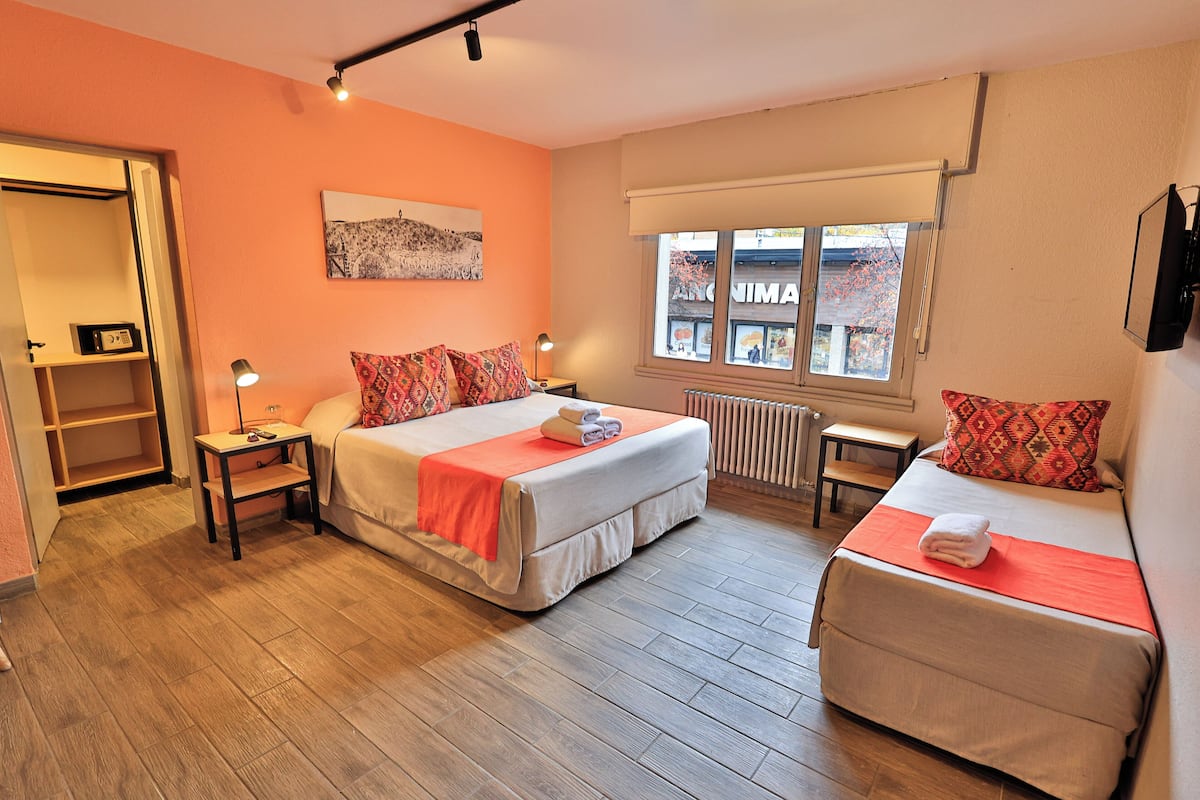 Une chambre d’hôtel avec deux lits, une fenêtre donnant sur un immeuble, un radiateur et une étagère avec une horloge.
