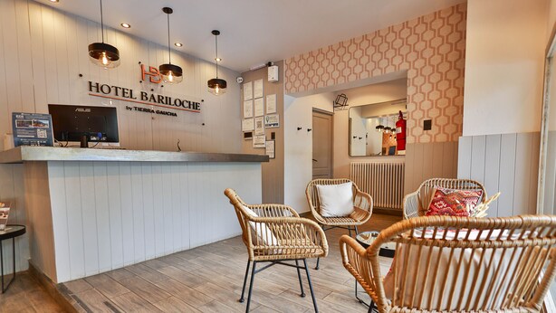 A hotel reception area with a wooden floor, wicker chairs, a reception desk with a computer, and a wall-mounted sign for Hotel Bariloche by Tierra Gaucha. - Hotel Bariloche By Tierra Gaucha (San Carlos de Bariloche)