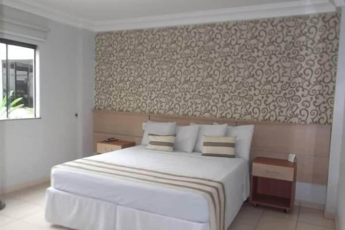 Une chambre à coucher avec un grand lit, une tête de lit recouverte d’un papier peint à motifs, une table de chevet et une fenêtre.