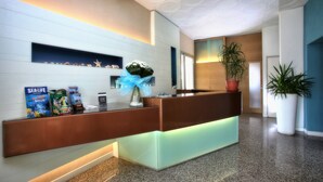 A reception desk with a modern design, featuring a glass partition and a wooden counter. There are promotional items and a vase with flowers on the desk. - Hotel Dante (Jesolo)