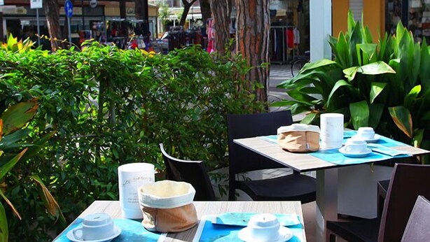 An outdoor café with tables set for two, each with a white cup, saucer, and a small bowl. - Hotel Dante (Jesolo)