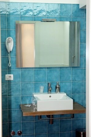 A bathroom with blue tiles, a white sink, a mirror, a telephone, and a towel dispenser. - Hotel Dante (Jesolo)