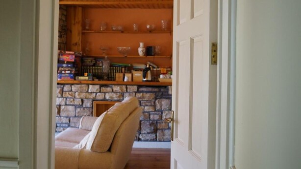 Un pasillo que conduce a una sala de estar con chimenea de piedra, un sofá y una zona de cocina con estantes de madera y diversos objetos.