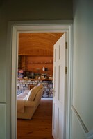 A hallway leading to a living room with a stone fireplace, a sofa, and a kitchen area with wooden shelves and various items.