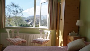 Un dormitorio con cama, dos sillas, una mesita de noche y ventana con vistas a árboles y colinas.