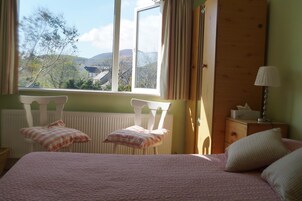 A bedroom with a bed, two chairs, a nightstand, and a window with a view of trees and hills.