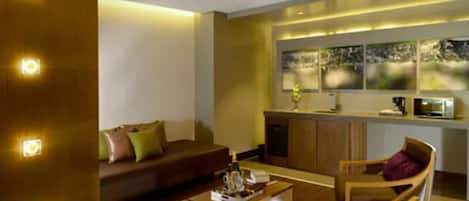 Una sala de estar moderna con un sofá, una mesa de centro y una silla de madera. Se ve un área de cocina con una barra y electrodomésticos al fondo.
