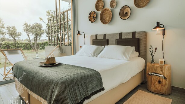 A bedroom with a large bed, a view of the outdoors, and a wooden nightstand with a lamp. - Herdade da Rocha Boutique Lodge (Crato)