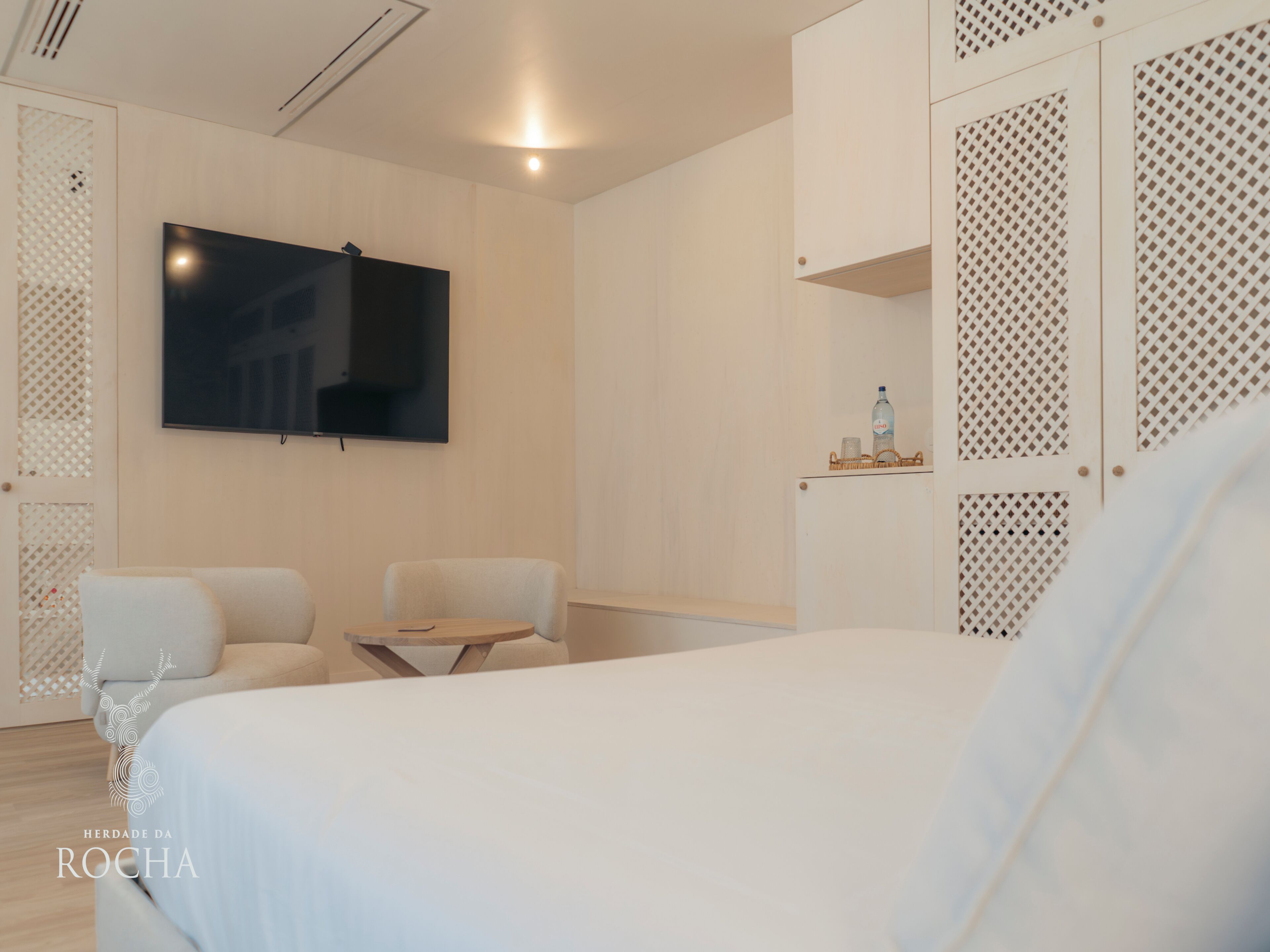 A modern hotel room with a large bed, a flat-screen TV mounted on the wall, a small table with a chair, and a decorative shelf with a bottle.