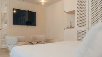 A modern hotel room with a large bed, a flat-screen TV mounted on the wall, a small table with a chair, and a decorative shelf with a bottle.