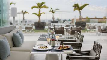 A dining area with tables and chairs, a sofa with cushions, and a view of palm trees and the ocean.