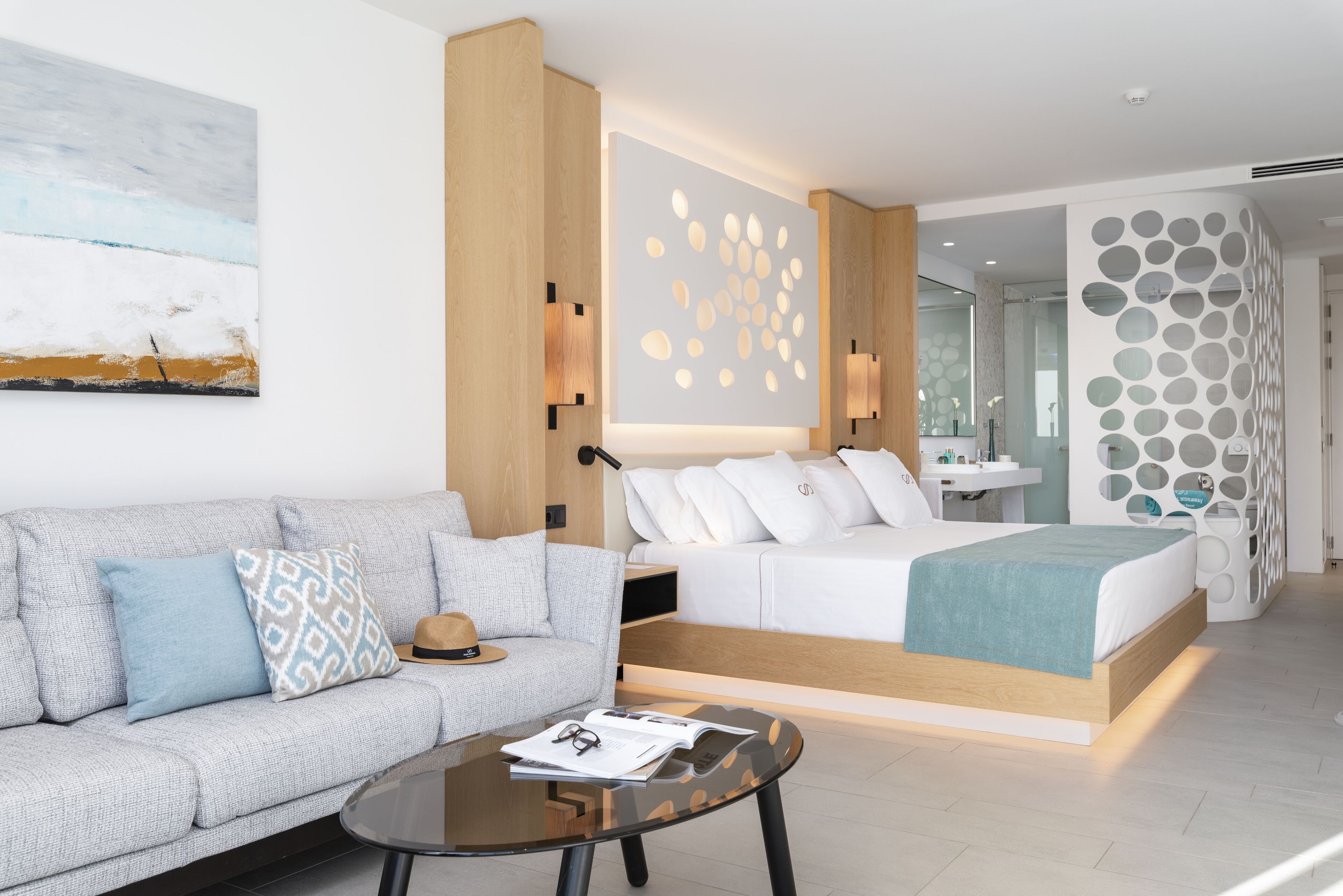 A modern hotel room with a sofa, a bed, a glass coffee table, and a wall with circular patterns.