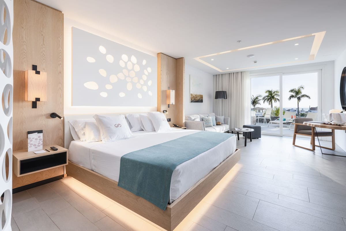 A modern hotel room with a large bed, a desk, a sofa, and a balcony with a view.