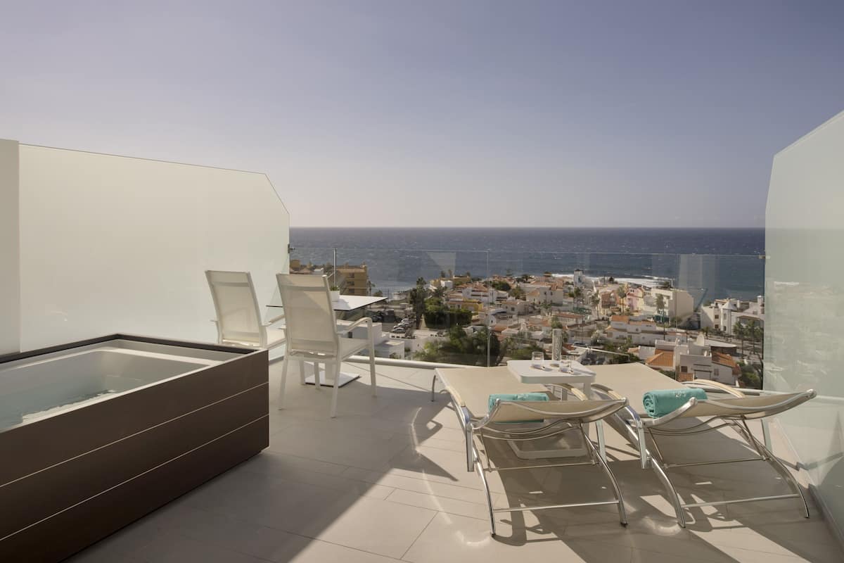 A balcony with a glass enclosure, white chairs, and a table overlooking a coastal cityscape.