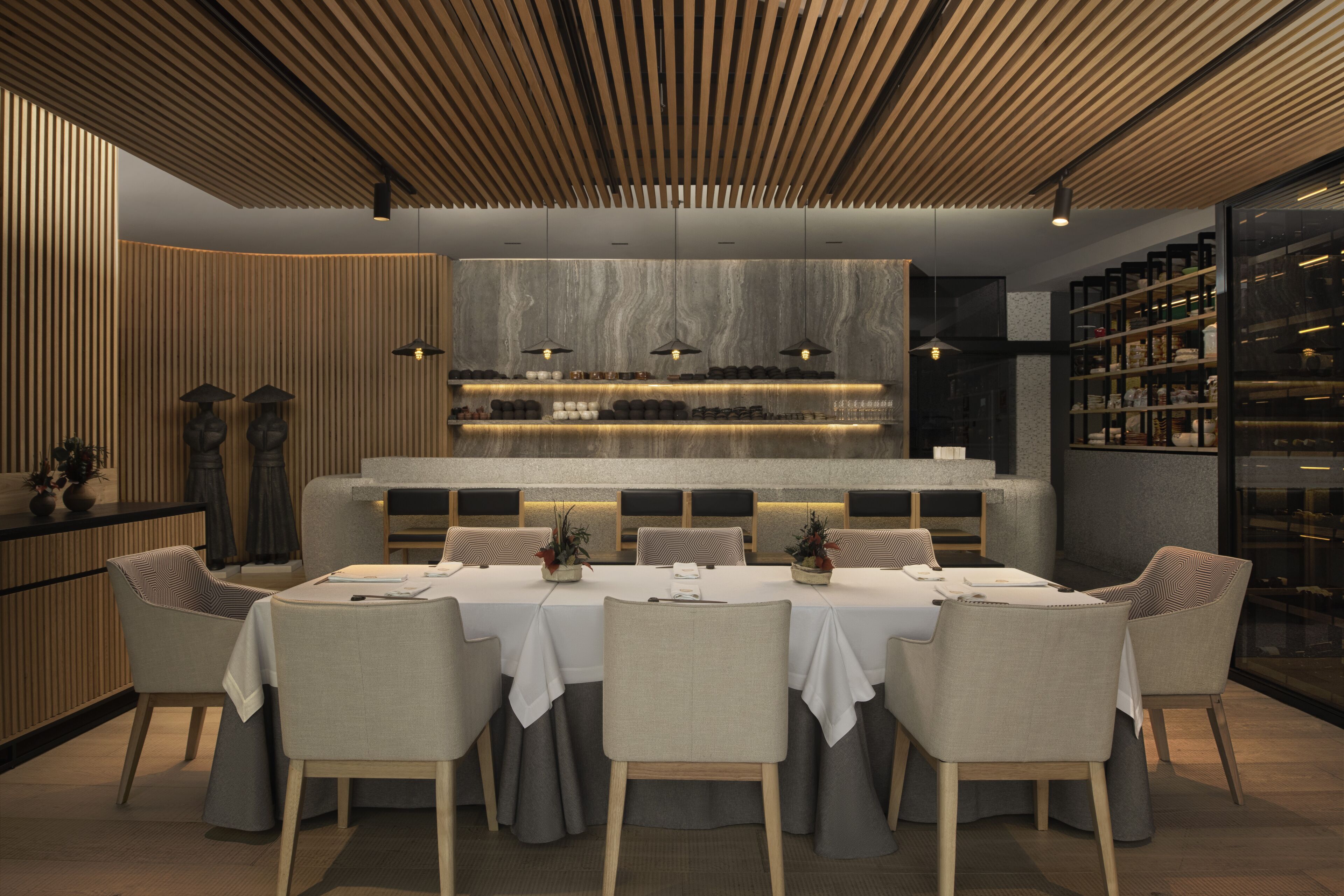 A modern dining area with a long table, cushioned chairs, and a central bar area with shelves.