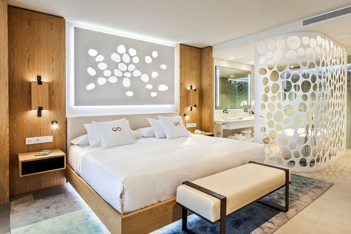 A modern bedroom with a large bed, bedside tables, a bench, a wall-mounted light, and a bathroom visible through a glass door.