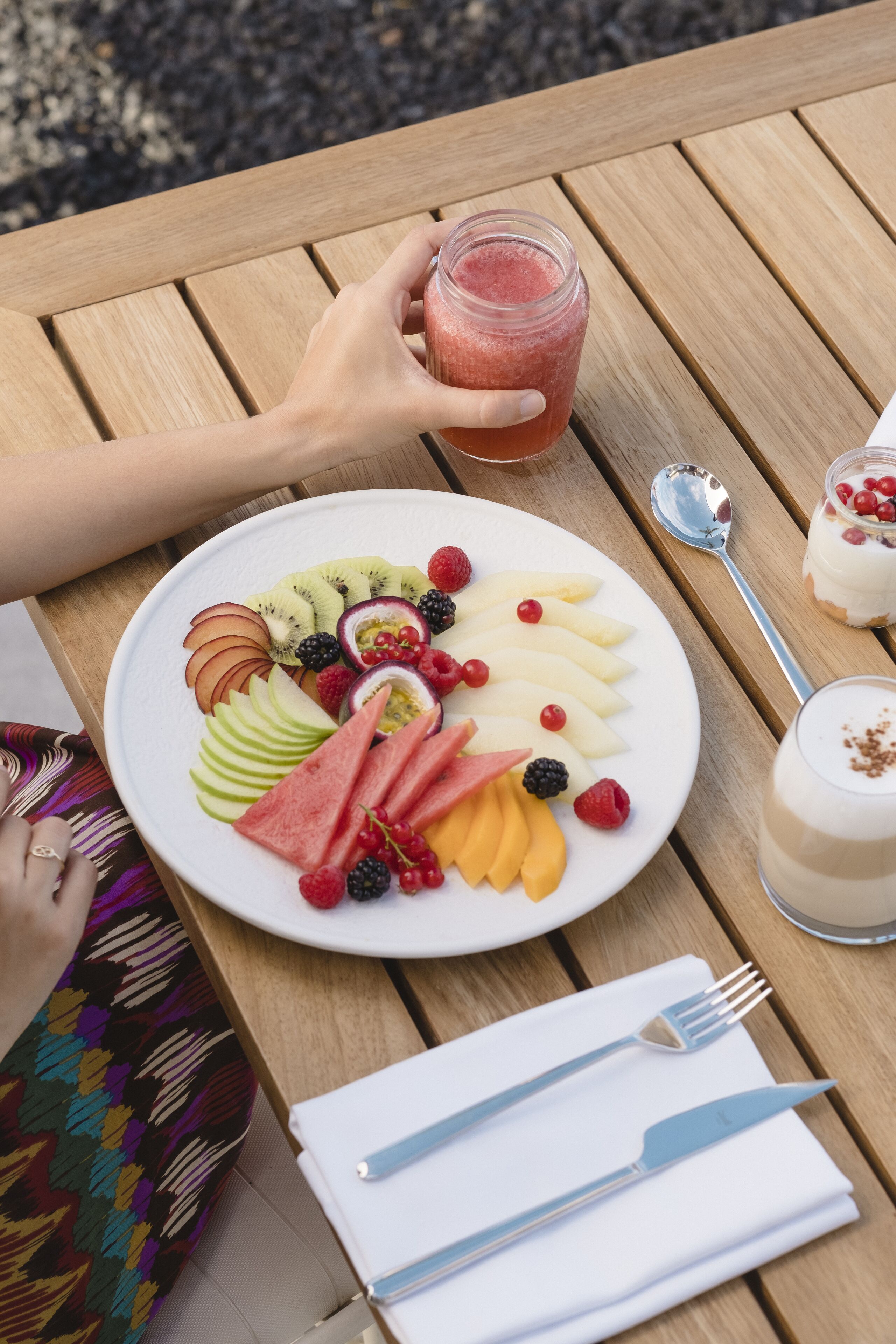 A wooden table with a plate of fruit, a glass of smoothie, and a dessert.