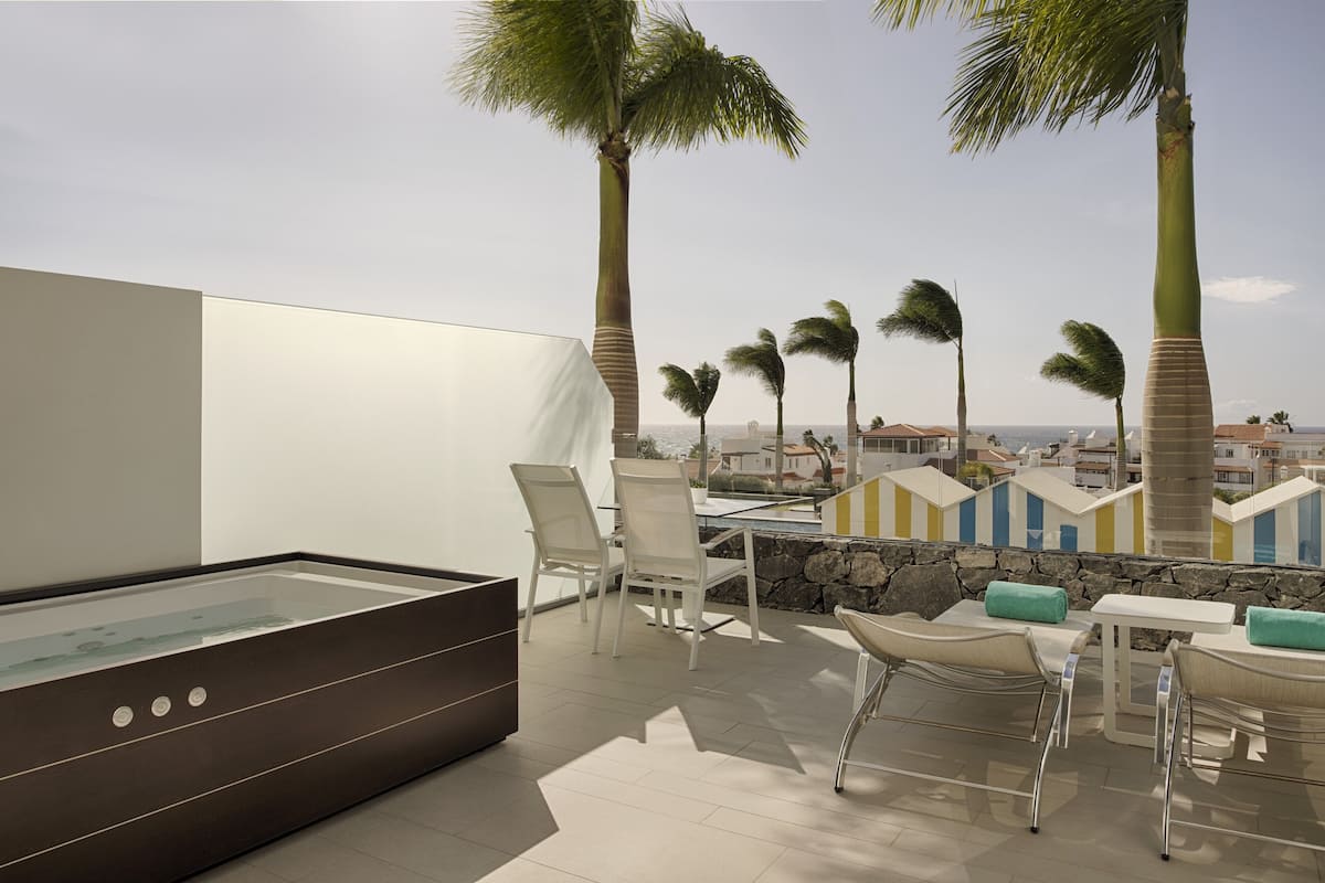 A modern outdoor terrace with a glass-enclosed hot tub, white chairs, and a stone wall.