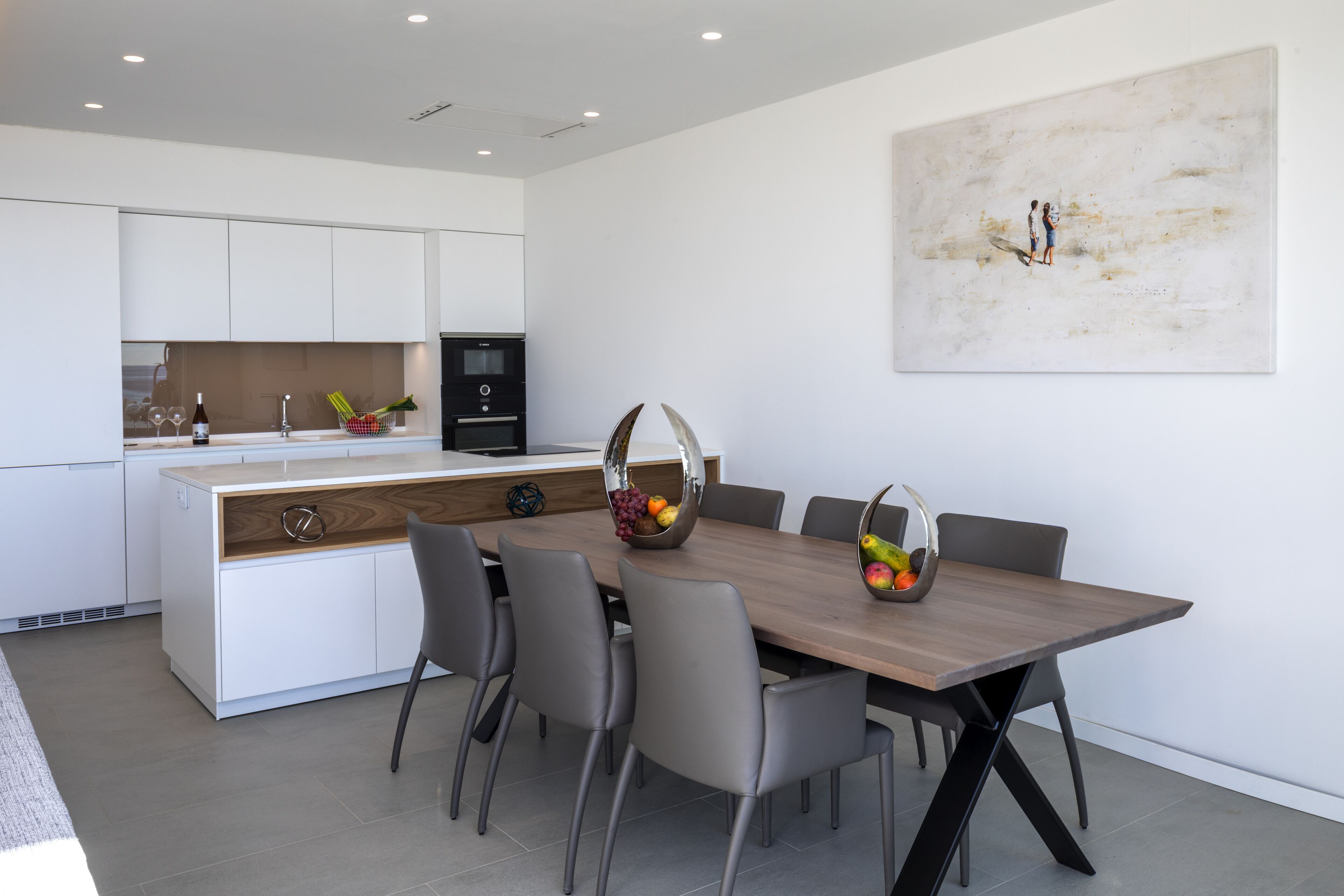 A modern kitchen with white cabinets, a wooden dining table, and a large abstract painting on the wall.