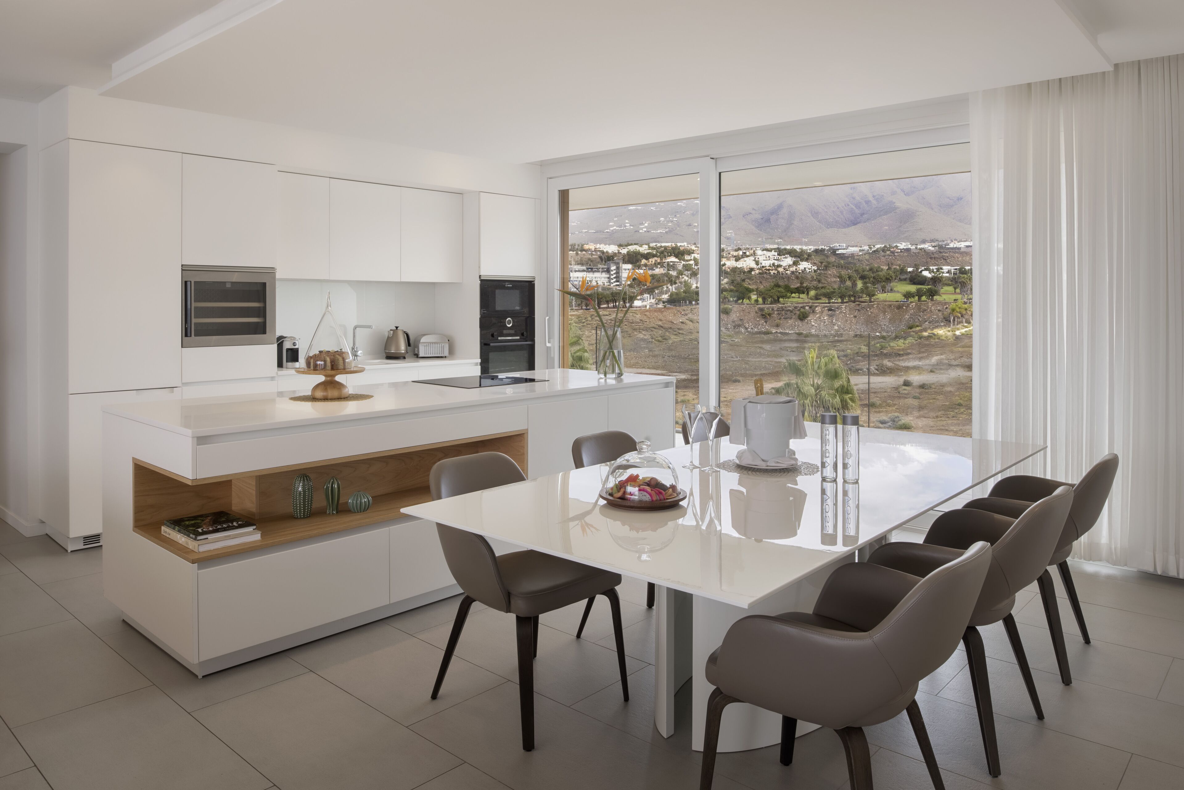 Una cocina moderna con armarios blancos, una isla central con asientos y un amplio ventanal con vistas a un paisaje montañoso.