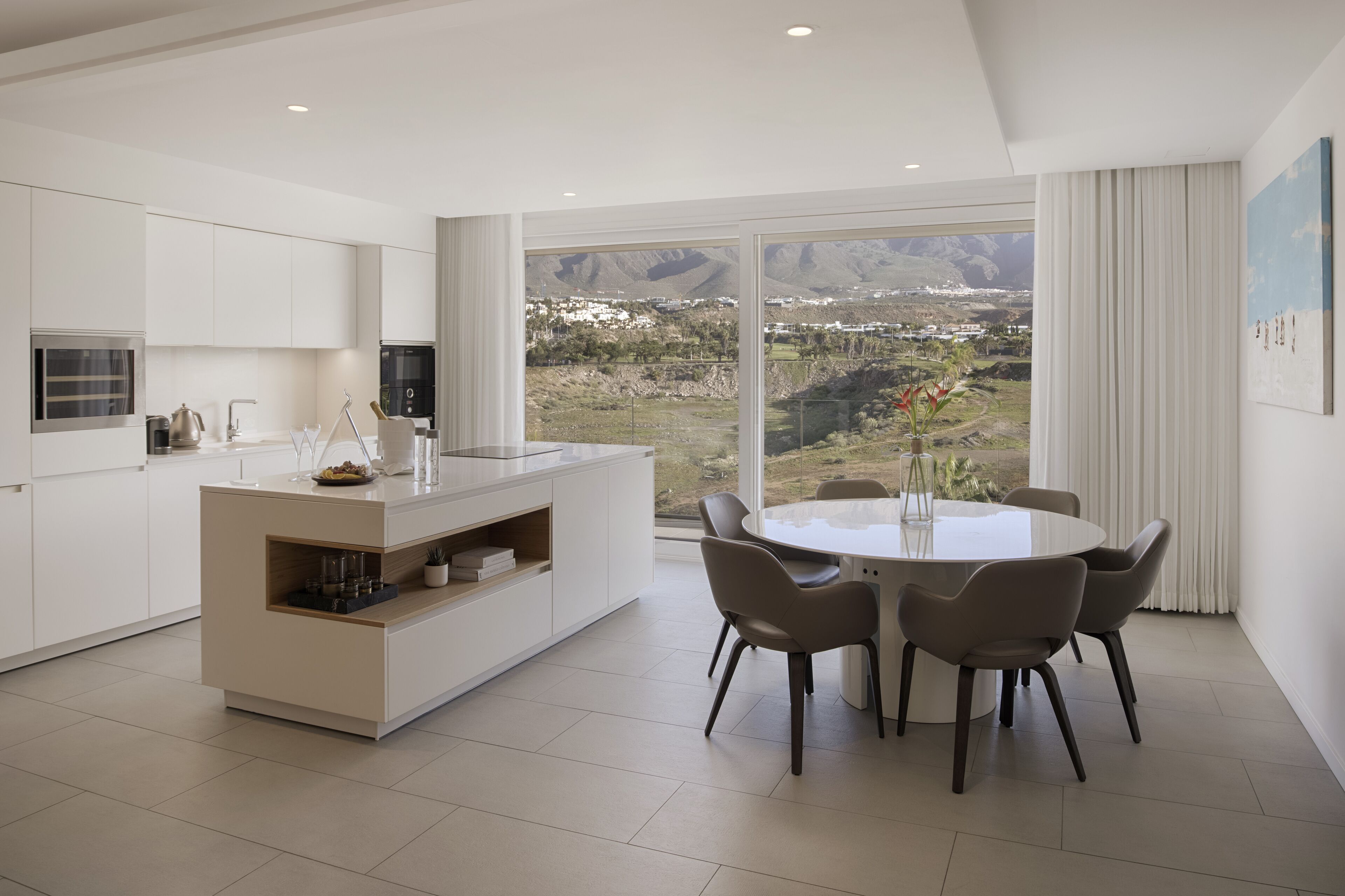 A modern kitchen with a central island, dining area with chairs, and a large window offering a view of a mountainous landscape.