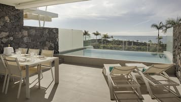 Un área de comedor moderna al aire libre con pared de piedra, piscina y vista al océano.