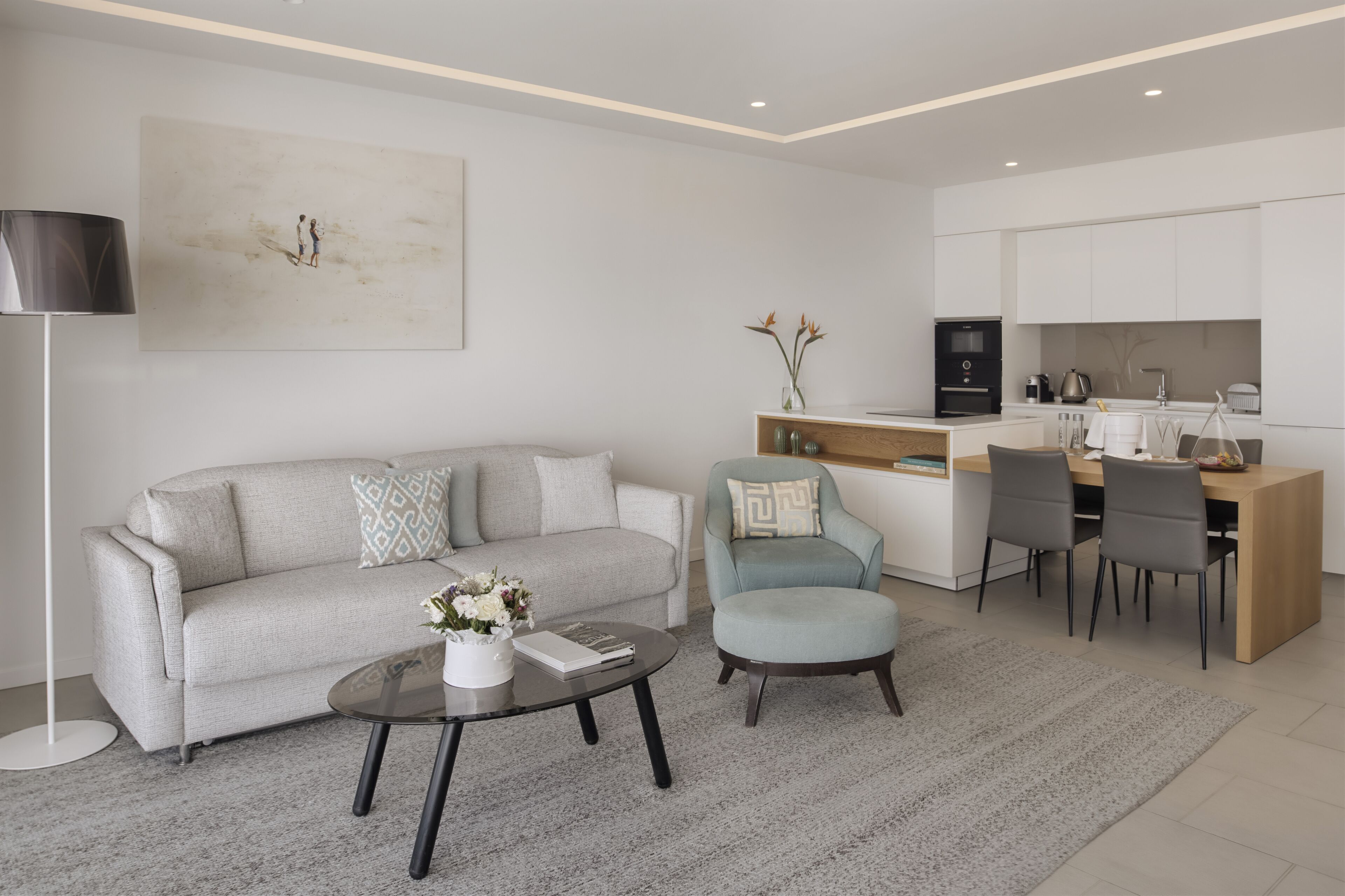 A modern living room with a sofa, a coffee table, and a dining area with chairs and a kitchen in the background.