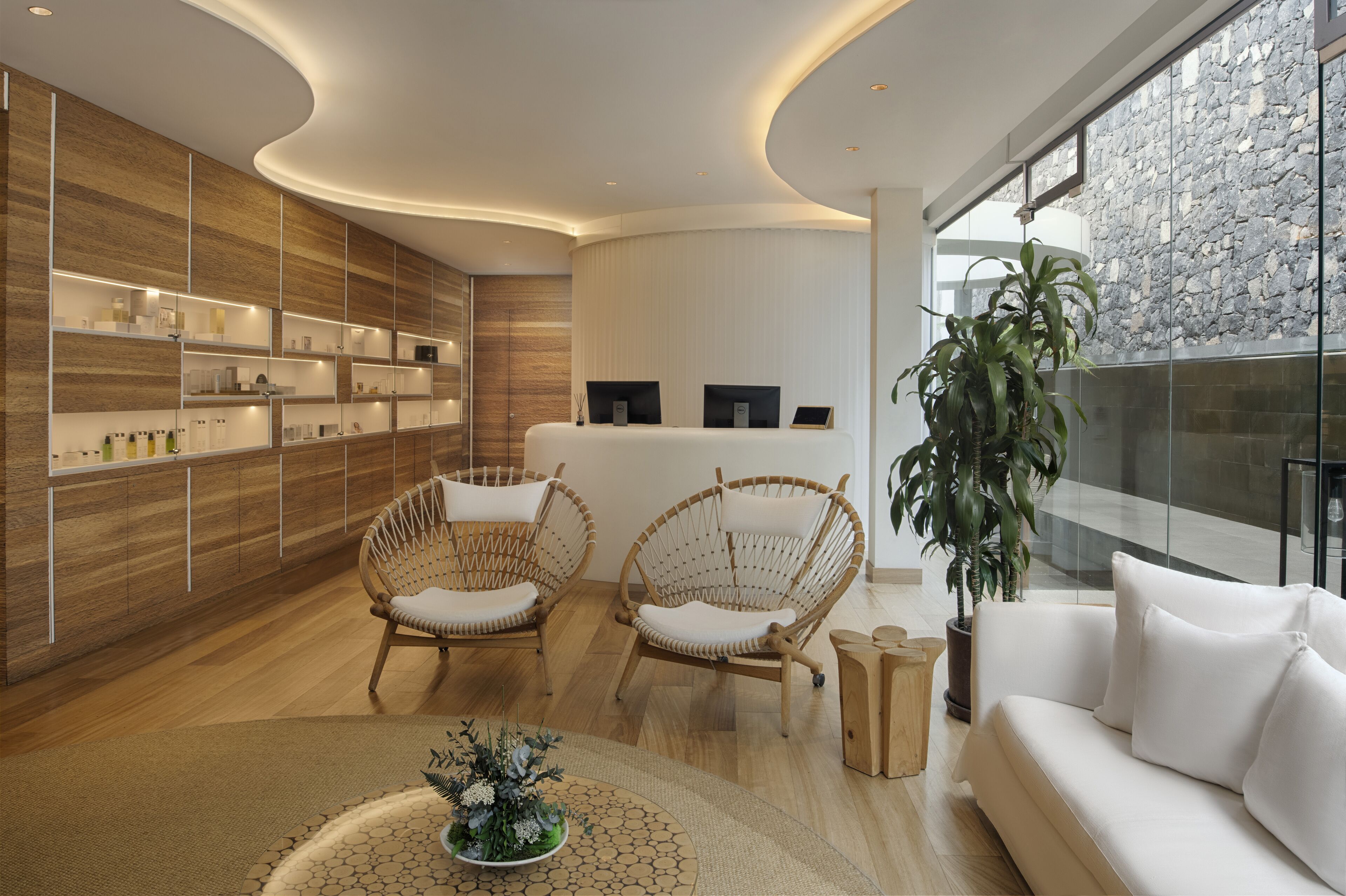 Un área de recepción moderna con paneles de madera, dos sillones de mimbre, un sofá blanco y un mostrador de recepción.