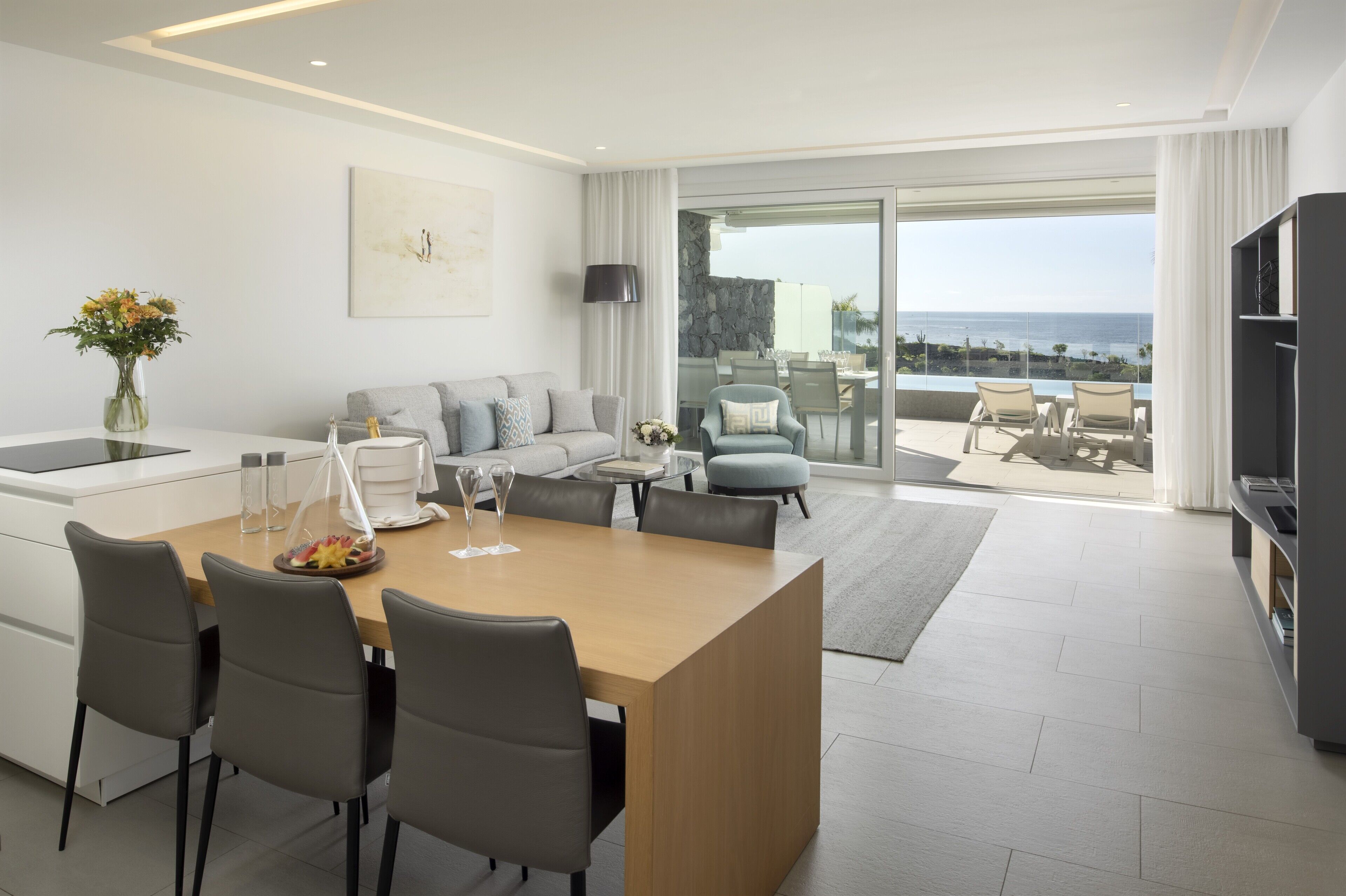 Una habitación de hotel moderna con comedor, un conjunto de sofás, televisión y un balcón con vista al océano.