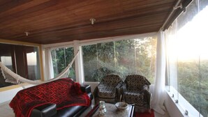 A room with a wooden ceiling, a hammock, a black sofa, patterned armchairs, and a red rug.
